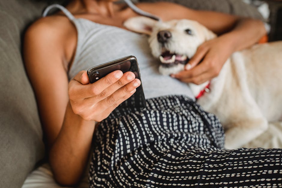 Faceless ethnic female with cellphone caressing dog with open mouth while resting on bed in house